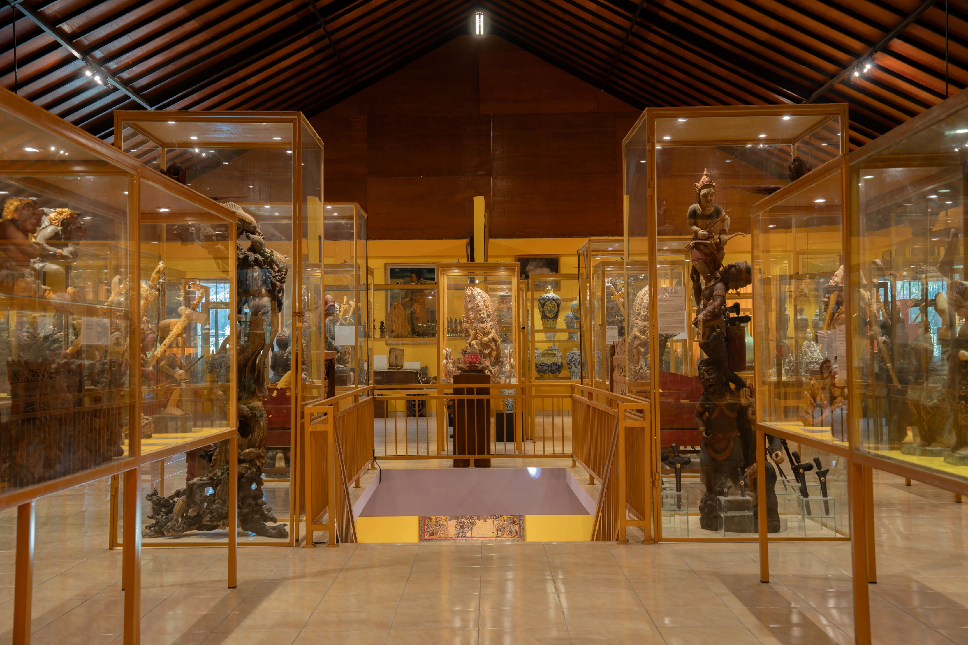 Interior gallery at Museum PBC featuring glass display cases with traditional Indonesian wood carvings, statues, and cultural artifacts under a vaulted wooden ceiling.